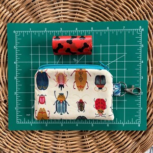 May include: A white zippered pouch with a teal zipper and a key ring. The pouch has a pattern of colourful beetles on it.