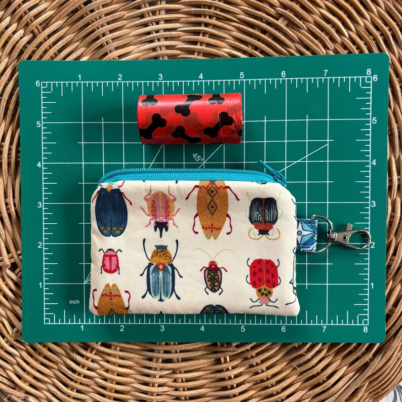 May include: A white zippered pouch with a teal zipper and a key ring. The pouch has a pattern of colourful beetles on it.