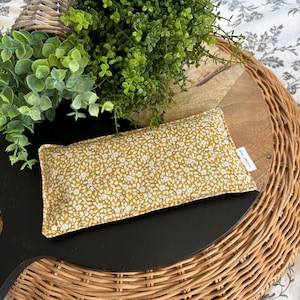 May include: A mustard-yellow fabric item with a white floral pattern. The item is rectangular and rests on a black surface, surrounded by greenery and a woven basket. The item has a tag that reads "October Sage".