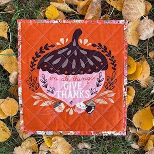 Peut inclure: Un carré de tissu orange matelassé avec un motif d'écorce de gland rose et marron. Le gland porte le texte "in all things give thanks" à l'intérieur. Le tissu est entouré d'une bordure florale.