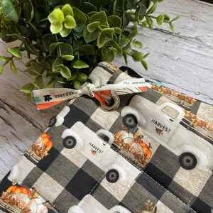 May include: A black and white checked fabric tote bag with a white truck design and the word "HARVEST" printed on the side. The truck is carrying pumpkins and autumn leaves. The bag has a black and white ribbon with the words "AND FREE" printed on it.