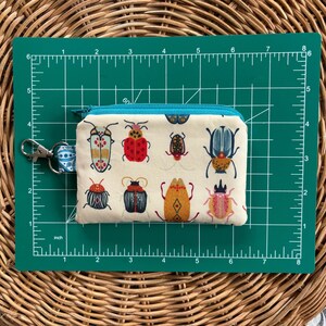 May include: A white fabric zipper pouch with a turquoise zipper and a colourful insect pattern. The pouch is 7.6 cm wide and 12.7 cm long.