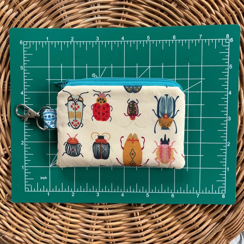 May include: A white fabric zipper pouch with a turquoise zipper and a colourful insect pattern. The pouch is 7.6 cm wide and 12.7 cm long.