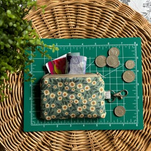 Peut inclure: Une petite pochette zippée verte avec un imprimé floral de fleurs blanches. La pochette est ouverte et contient de l'argent liquide et des pièces de monnaie. La pochette est posée sur un tapis de coupe vert avec une règle.