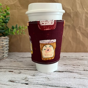 May include: A burgundy fabric coffee cup sleeve with a pattern of jars with labels that say "sweet jam" and "honey".