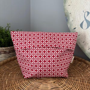 May include: A red and white patterned fabric pouch. The pouch is rectangular with a folded-over top. The pattern is a repeating design of red crosses on a white background. The pouch is sitting on a woven wicker tray.