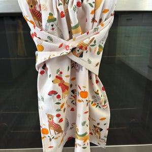 May include: White kitchen towel featuring a pattern of dogs in hats and scarves, pumpkins, mushrooms, and autumn leaves. The towel is looped over a stainless steel oven handle.