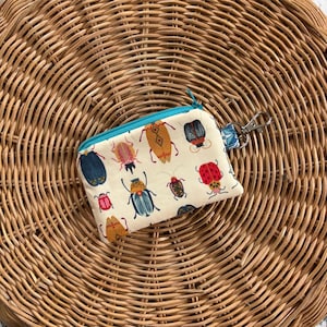 May include: A small white zippered pouch with a teal zipper and a silver key ring. The pouch is decorated with a colourful pattern of various insects, including beetles, butterflies, and ladybugs.