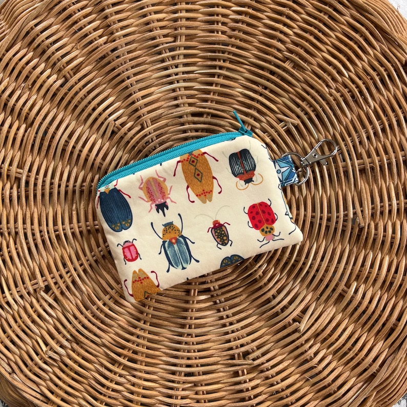 May include: A small white zippered pouch with a teal zipper and a silver key ring. The pouch is decorated with a colourful pattern of various insects, including beetles, butterflies, and ladybugs.