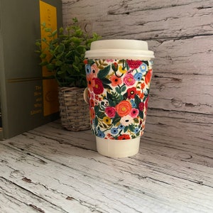 May include: A white coffee cup with a floral print sleeve. The sleeve has a white background with red, orange, pink, yellow, blue, and green flowers. The cup is sitting on a wooden surface.