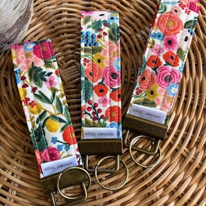 May include: Three floral key fobs with brass fittings. The fabric displays a colourful pattern of red, pink, yellow, and blue flowers on a white backdrop. Each fob has a small label that reads "October Days".