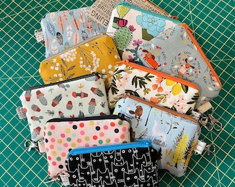 May include: A collection of nine colourful zippered pouches with various patterns and designs. The pouches are made of fabric and have a variety of colours, including blue, green, red, yellow, and white. The pouches are decorated with different patterns, including floral, geometric, and abstract designs. Some of the pouches have text printed on them, including the words "Zurich", "London", "Jakarta", and "Lisbon".
