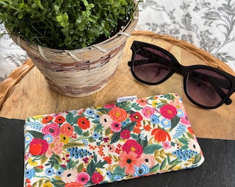 May include: A glasses case with a vibrant floral print featuring red, pink, and blue flowers on a white base. Black sunglasses are placed on a wooden tray. A small potted plant is also visible in the image.