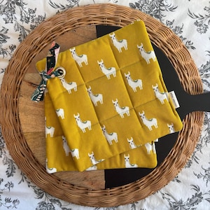 May include: Two yellow quilted pot holders with a white llama pattern. The pot holders are on a woven wicker surface with a black trivet.