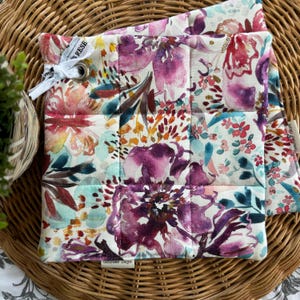 Peut inclure: Deux maniques matelassées avec un motif floral dans des tons de rose, violet, turquoise et jaune. Les maniques ont un fond blanc et sont matelassées en forme de carré. Le texte "October Days" est imprimé sur l'une des maniques.