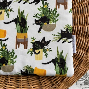 May include: White fabric featuring a repeating pattern of black cats amongst yellow and brown plant pots with green foliage. The cats have yellow eyes. The text "October Days" is on a white label.
