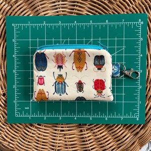 May include: A small white zippered pouch with a colourful insect print. The pouch has a blue zipper and a silver key chain attached.