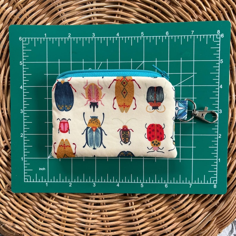 May include: A small white zippered pouch with a colourful insect print. The pouch has a blue zipper and a silver key chain attached.