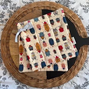 May include: Two beige pot holders with a pattern of colourful beetles on a woven wicker surface. The pot holders have a loop for hanging.