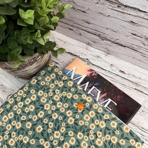 May include: A green book cover with a floral pattern of small white flowers and dark green leaves. The cover is secured with a small orange button. A book with the title "MAEV" is partially visible.