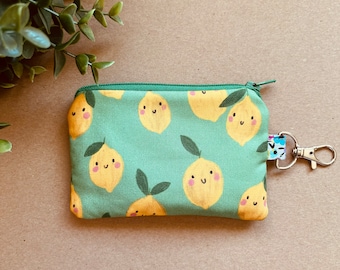 May include: A green zippered pouch with a lemon print. The lemons have smiling faces and are yellow with green leaves. The pouch has a silver key ring attached.