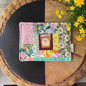 May include: A patchwork fabric item with a honey jar design. The item features a variety of floral patterns in pink, blue, green, and yellow. The honey jar is centered on a burgundy background, with the word "Honey" written on it.