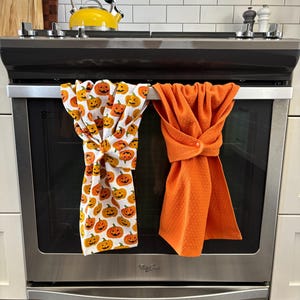 Peut inclure: Deux torchons de cuisine suspendus à un four. L'un est blanc avec des motifs de citrouilles oranges, l'autre est orange uni. Le four est en acier inoxydable avec un dessus noir, et une bouilloire jaune est visible.