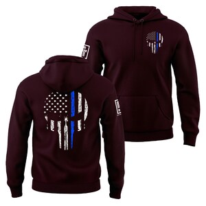 May include: Burgundy hoodie featuring a skull design with an American flag and a blue line. The design is on the front and back. The hoodie has a hood and a front pocket. The sleeves have a small white logo.