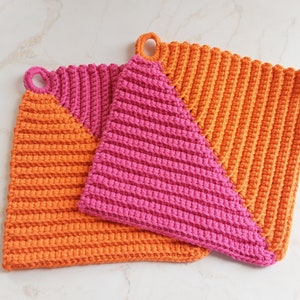 Könnte beinhalten: Zwei gehäkelte Topflappen, einer orange und einer pink, mit einem diagonalen Split-Design. Beide Topflappen haben eine Schlaufe zum Aufhängen.