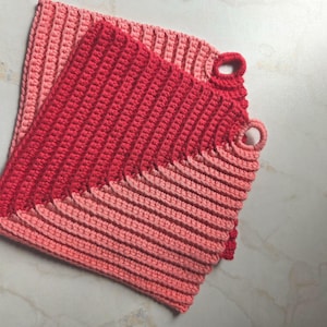 Könnte beinhalten: Zwei handgefertigte Topflappen aus Häkelarbeit in Rot- und Rosatönen. Die Topflappen haben eine gerippte Textur und eine Schlaufe zum Aufhängen. Der obere Topflappen ist dunkler rot, während der untere heller rosa ist.