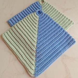 Op de afbeelding: Twee gehaakte ovenwanten, een lichtgroen en een lichtblauw, met een diagonale naad in het midden. Beide ovenwanten hebben een lus aan de bovenkant.