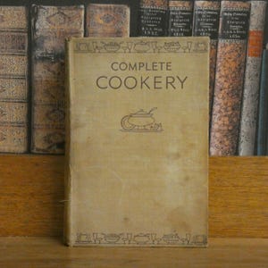 Könnte beinhalten: Ein Vintage-Kochbuch mit dem Titel "COMPLETE COOKERY" und einem beigefarbenen Einband. Der Titel ist in einer Serifenschrift gehalten, zusammen mit einer Grafik eines Kochtopfs. Das Buch steht auf einem Holzregal, mit anderen Büchern im Hintergrund.