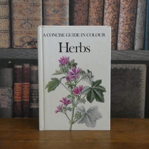 Op de afbeelding: Een wit boek met de titel "Herbs" en de tekst "A Concise Guide in Colour". De omslag toont een botanische illustratie van een plant met roze bloemen en groene bladeren. Het boek ligt op een houten oppervlak.