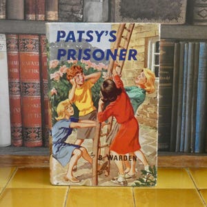 Op de afbeelding: Een vintage boek getiteld "Patsy's Prisoner" van B. Warden. De omslagillustratie toont vier jonge mensen bij een ladder, met een gebouw en bloemen op de achtergrond. Het boek staat tegen een achtergrond van een boekenplank.
