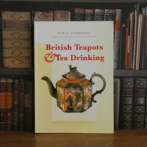 May include: A book titled "British Teapots & Tea Drinking" by Robin Emmerson. The cover features a detailed illustration of a decorative teapot with green, orange, and white accents. The book is set against a backdrop of antique books.