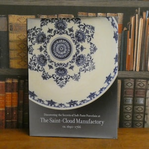 May include: A book cover featuring a white plate with a blue floral design. The plate is partially visible, with the rest of the cover displaying text: "Discovering the Secrets of Soft-Paste Porcelain at The Saint-Cloud Manufactory ca. 1690-1766."