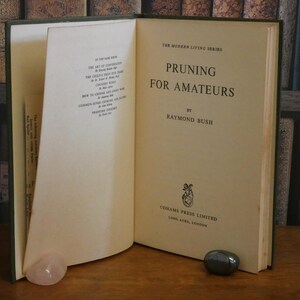 May include: An open book titled "Pruning for Amateurs" by Raymond Bush. The book is part of "The Modern Living Series." The cover is green, and the pages are off-white. A pink and a dark gray stone are at the base of the book.