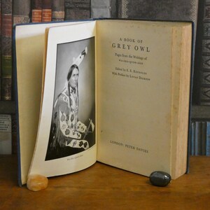 Könnte beinhalten: Ein offenes Buch mit dem Titel "A Book of Grey Owl" mit einem Schwarz-Weiß-Porträt eines amerikanischen Ureinwohners. Das Buch hat einen blauen Einband und liegt auf einer Holzoberfläche mit zwei kleinen Steinen.
