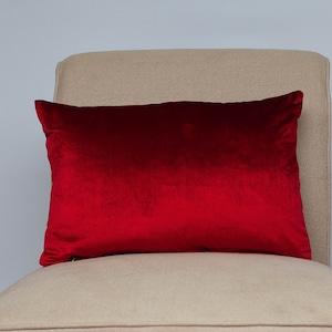 Op de afbeelding: Een rood fluwelen kussen op een beige stoel. Het kussen is rechthoekig en heeft een gladde, zachte textuur.