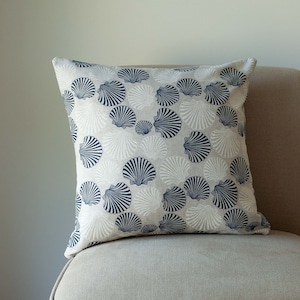 May include: Square cushion with a seashell pattern. The cushion has a light grey background with a repeating pattern of navy blue and white seashells. It is placed on a beige chair.
