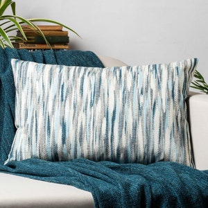 May include: A rectangular throw pillow with a blue and white abstract pattern. The pillow is on a white sofa with a teal throw blanket.