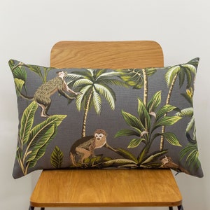 May include: Rectangular cushion with a grey background depicting a jungle scene. The design features green palm trees, lush foliage, and monkeys in brown and tan hues. The cushion is placed on a wooden chair.