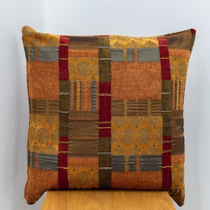 Può includere: Un cuscino decorativo con un design a patchwork in varie tonalità di marrone, oro e rosso. Il cuscino ha un motivo geometrico con un sottile design a spirale.