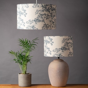 Könnte beinhalten: Zwei Lampen mit cremefarbenen Schirmen, die mit einem blauen Blumenmuster verziert sind. Die größere Lampe hängt von der Decke, während die kleinere Lampe auf einem Tisch mit einem braunen Keramikfuß steht. Im Vordergrund steht eine grüne Topfpflanze.