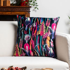 Pode incluir: Almofada quadrada com um padrão floral vibrante. A almofada apresenta um fundo preto com ilustrações botânicas coloridas em tons de rosa, amarelo, azul e verde. A almofada está colocada num sofá branco.