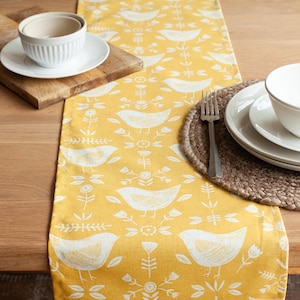 Op de afbeelding: Een gele tafelrunner met een wit vogelpatroon. De runner ligt op een houten tafel met een gedekte tafel met een witte kom en een bord.