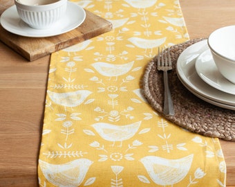 May include: A yellow table runner with a white bird pattern. The runner is on a wooden table with a place setting of a white bowl and a plate.