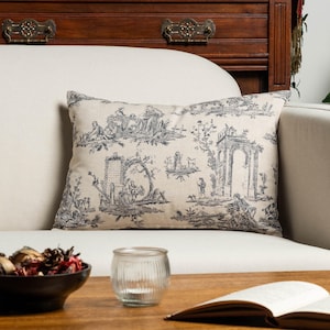 May include: A rectangular throw pillow featuring a beige fabric base with a dark grey toile de jouy pattern. The print depicts detailed pastoral scenes with figures, trees, and architectural ruins, including a prominent stone archway structure.