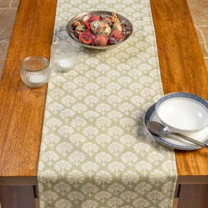 May include: A sage green table runner featuring a repeating white floral design. The runner is laid on a wooden table, with a bowl of decorative items, candles, and a bowl with a spoon and plate.