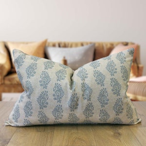 May include: A rectangular cushion with a light beige fabric, featuring a repeating pattern of blue floral motifs. The cushion rests on a wooden surface, with a blurred background showing a sofa and other cushions.